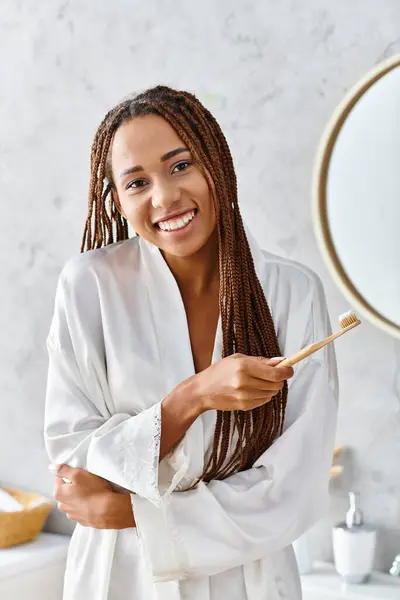 Афроамериканка в халате с афрокосичками чистит зубы в современном зеркале в ванной. — стоковое фото