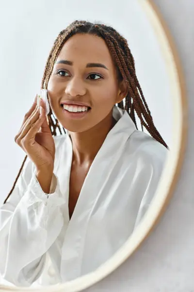 Uma mulher afro-americana com tranças afro usando almofada de algodão com toner na frente de um espelho em um banheiro moderno, enquanto vestindo um roupão de banho. — Fotografia de Stock
