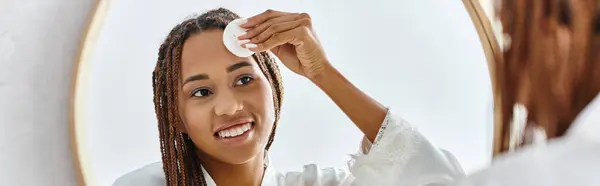 Une Afro-Américaine avec des tresses afro en coton avec du toner devant un miroir dans une salle de bain moderne, mettant l'accent sur la beauté et l'hygiène. — Photo de stock