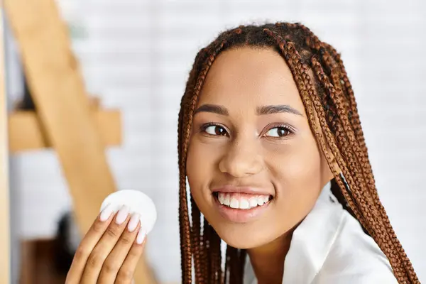 Femme afro-américaine avec des tresses en peignoir, tenant un objet blanc dans sa main dans une salle de bain moderne. — Photo de stock