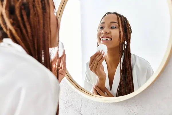 Uma mulher Africano-Americana com tranças afro em um roupão de banho usando almofada de algodão na frente de um espelho em um banheiro moderno. — Fotografia de Stock