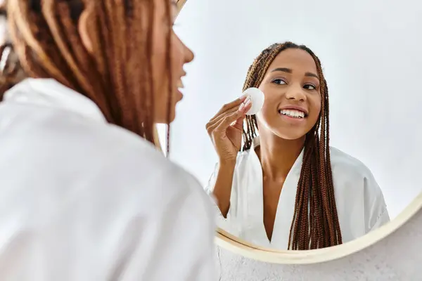 Uma mulher afro-americana com tranças afro usando almofada de algodão na frente de um espelho em um banheiro moderno, com foco em beleza e higiene. — Fotografia de Stock