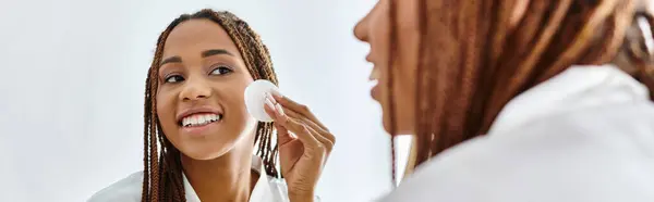 An African American woman in a bathrobe using cotton pad while looking at herself in the mirror of a modern bathroom. — Stock Photo