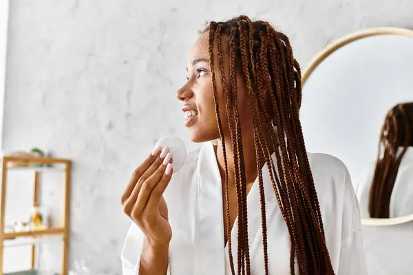 Africano mulher americana com tranças afro fica na frente do espelho em roupão de banho, segurando almofada de algodão no banheiro moderno. — Fotografia de Stock