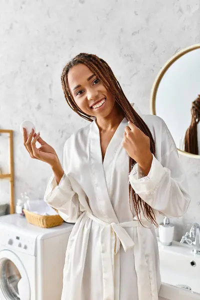 Афроамериканка с афрокосичками в халате, держащая хлопковую палочку в современной ванной, сосредоточенная на красоте и гигиене. — стоковое фото