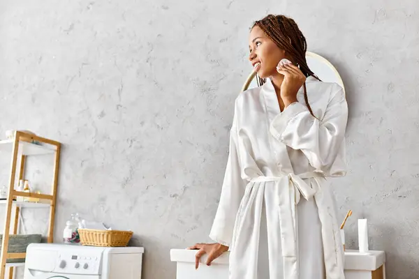 Una mujer afroamericana con trenzas afro, en un albornoz, sosteniendo una almohadilla de algodón en un baño moderno. - foto de stock