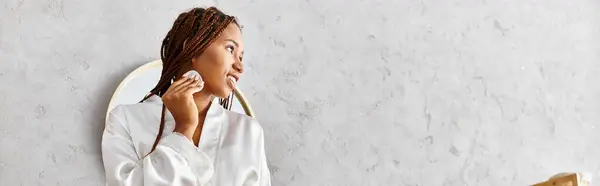African American woman with afro braids in bathrobe holding cotton pad in front of a mirror in a modern bathroom. — Stock Photo