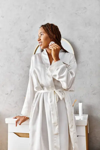 An African American woman with afro braids in a bathrobe holding cotton pad in a modern bathroom. — Stock Photo