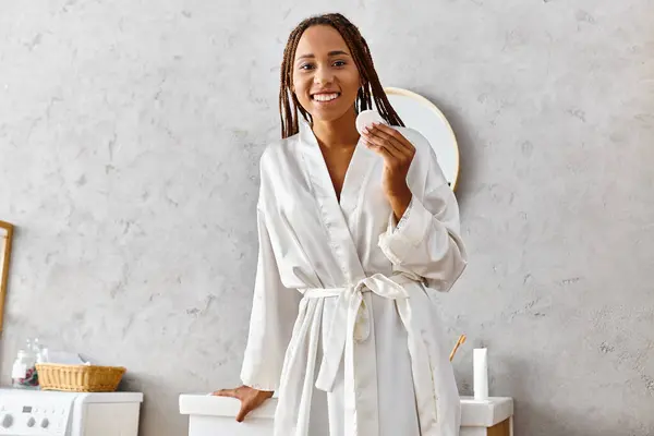 Une femme afro-américaine en robe blanche se tient gracieusement dans sa salle de bain moderne, rayonnant de beauté et de tranquillité. — Photo de stock