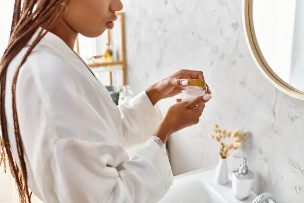 Afroamerikanerin mit Afro-Zöpfen in weißer Robe hält Schönheitsglas mit Creme im modernen Badezimmer für Schönheit und Hygiene. — Stockfoto