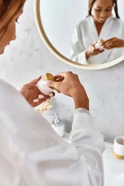 An African American woman with afro braids in a bathrobe holding beauty jar with cream in front of a mirror in a modern bathroom. — Stock Photo