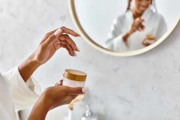 Una mujer afroamericana en un albornoz sosteniendo un frasco de crema frente a un espejo en su moderno baño. — Stock Photo