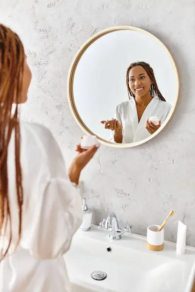 Eine afroamerikanische Frau mit Afro-Zöpfen steht in einem modernen Badezimmer und hält ein Schönheitsglas mit Creme in der Hand — Stockfoto