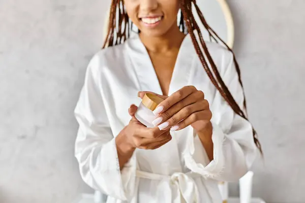 Uma mulher afro-americana com tranças afro fica em um banheiro moderno, segurando frasco de beleza com creme — Fotografia de Stock