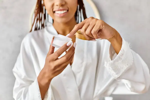 Una donna afroamericana vestita di bianco tiene delicatamente un barattolo di bellezza con crema nel suo bagno moderno. — Foto stock