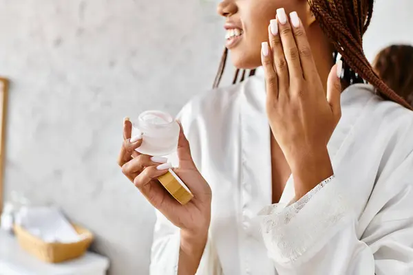 Femme afro-américaine avec des tresses en peignoir tenant un pot de crème devant son visage dans une salle de bain moderne. — Photo de stock