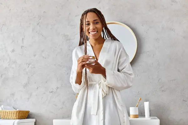 Une femme afro-américaine se tient devant un miroir, tenant un pot de beauté avec de la crème — Photo de stock