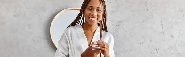 A stylish woman with dreadlocks holds a glass jar in front of a mirror in a modern bathroom, exuding beauty and confidence. — Stock Photo