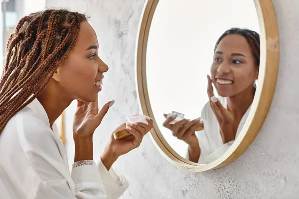 Una donna afroamericana con trecce afro applicare la crema in un bagno moderno mentre indossa un accappatoio. — Foto stock