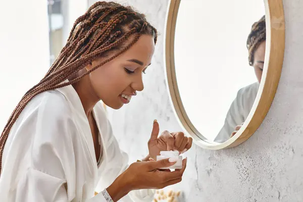 Femme afro-américaine en robe blanche tenant pot de beauté avec crème dans la salle de bain moderne avec des produits de beauté et d'hygiène. — Photo de stock