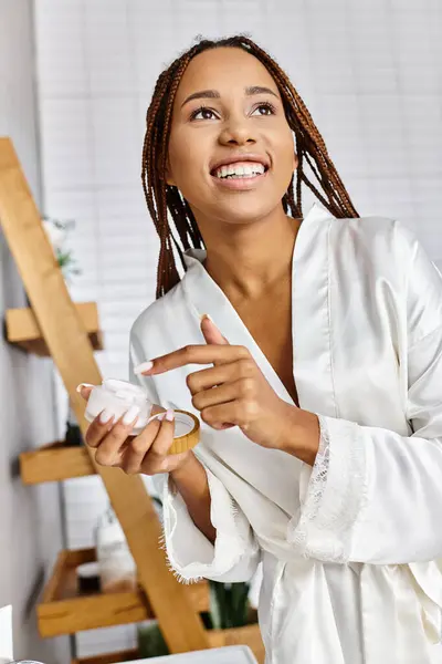Uma mulher afro-americana com tranças em um roupão de banho segurando frasco de beleza com creme — Fotografia de Stock