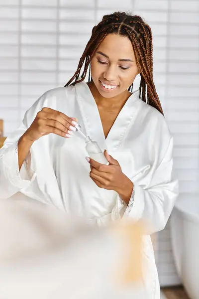 Eine afroamerikanische Frau mit Afro-Zöpfen im Bademantel hält eine Flasche Serum in einem modernen Badezimmer. — Stockfoto