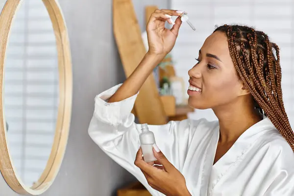African American woman with afro braids holding a bottle with serum in front of modern bathroom mirror. Beauty and hygiene routine. — Stock Photo