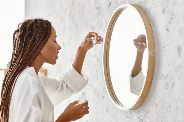 An African American woman with Afro braids in a bathrobe applying serum in a modern bathroom. — Stock Photo
