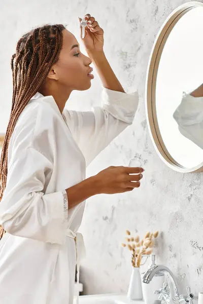 Une Afro-Américaine avec des tresses afro tenant une bouteille avec du sérum dans une salle de bain moderne tout en portant un peignoir de bain. — Photo de stock