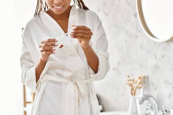Une femme afro-américaine joyeuse en peignoir, souriante et tenant un sérum dans une salle de bain moderne. — Photo de stock