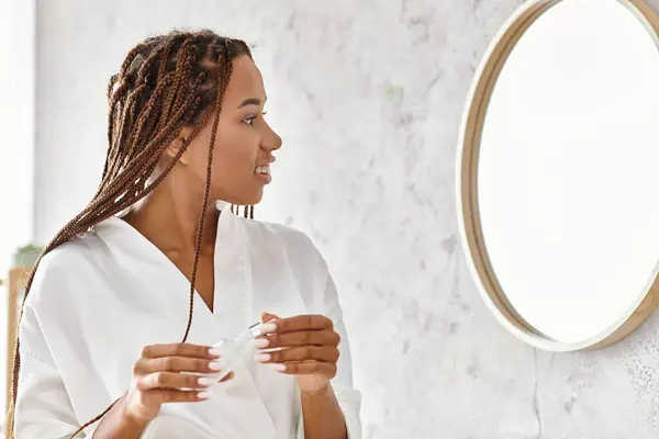 Africano mulher americana com dreadlocks em um roupão de banho, admirando-se no espelho de seu banheiro moderno. — Fotografia de Stock