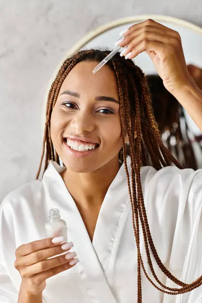 Una mujer afroamericana con trenzas afro en un albornoz se cepilla el pelo con gracia en un baño moderno. - foto de stock