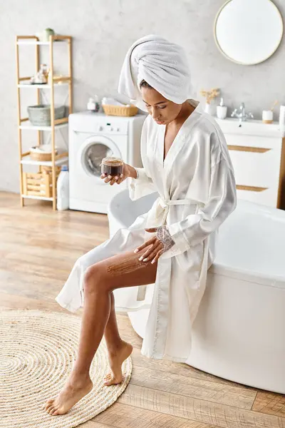 An African American woman in a bathrobe, exfoliating body with scrub while sitting in a bathtub in her modern bathroom. — Stock Photo