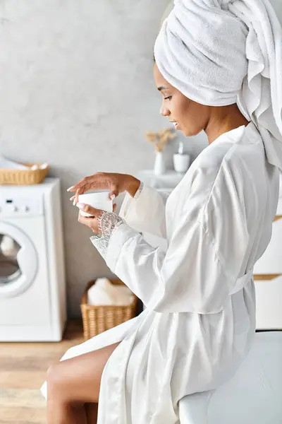 An African American woman with afro braids sits in a bathrobe and holding jar with cream in her modern bathroom. — Stock Photo