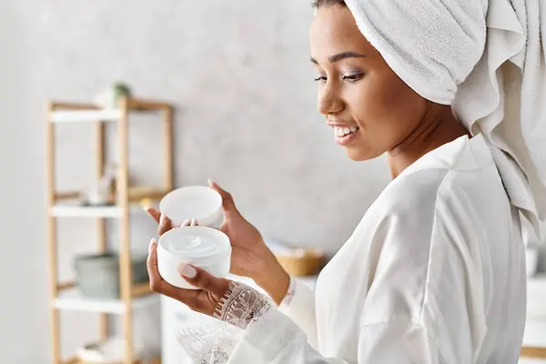Afroamerikanerin im Bademantel hält Container mit Handtuch auf dem Kopf im modernen Badezimmer. Schönheit und Hygiene-Routine. — Stockfoto
