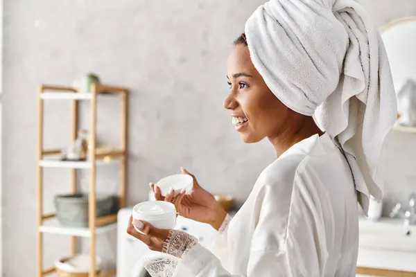 Una mujer, con una toalla en la cabeza, sosteniendo un frasco con crema en el baño, disfrutando de un momento de relajación y tranquilidad. - foto de stock