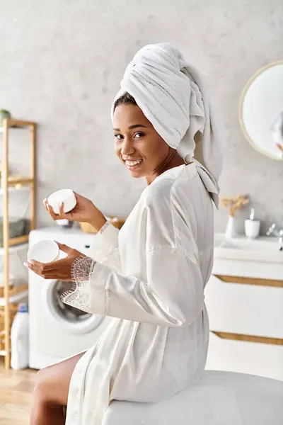 Una mujer afroamericana en un albornoz con una toalla envuelta alrededor de su cabeza, sosteniendo el frasco con crema - foto de stock