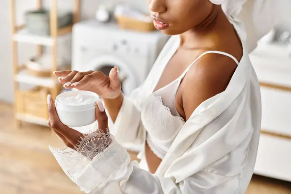 Africano mulher americana com tranças afro em vestido branco segurando frasco de creme no banheiro moderno. — Fotografia de Stock