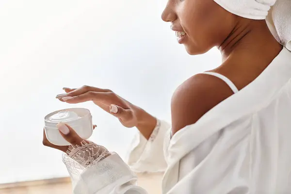 Афроамериканка в белом платье мирно держит банку крема в современной ванной комнате, олицетворяя красоту и элегантность. — стоковое фото