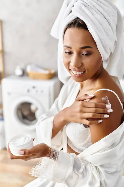 African American woman with afro braids in a bathrobe, towel on head, holding a jar of cream in her modern bathroom. — Stock Photo