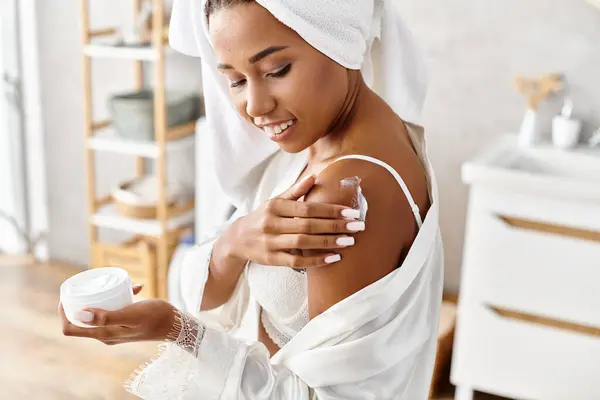 Femme afro-américaine en peignoir avec des tresses afro tenant pot de crème dans la salle de bain moderne. — Photo de stock