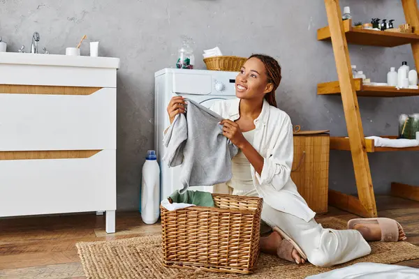 Uma mulher afro-americana com tranças afro senta-se ao lado de uma máquina de lavar roupa, no meio de lavar roupa em um banheiro. — Fotografia de Stock