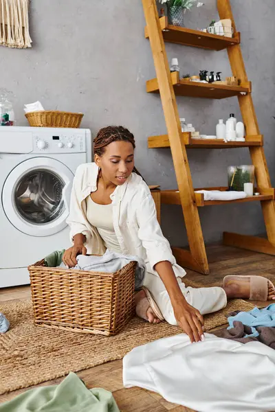 An African American woman with afro braids sits on the floor next to a laundry basket — Stock Photo