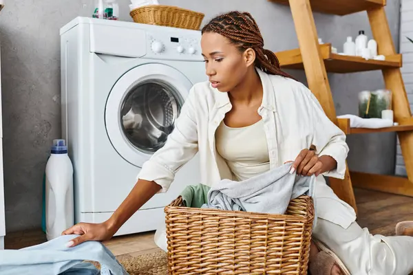 Une Afro-Américaine avec des tresses afro est assise sur le sol à côté d'une machine à laver, faisant la lessive dans un cadre de salle de bain. — Photo de stock