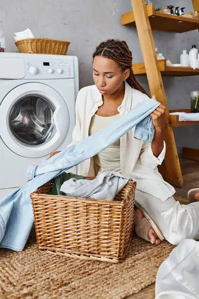 African American woman with afro braids doing laundry in a bathroom, sitting on the floor next to a washing machine. — Stock Photo