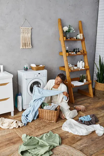 Une femme avec des tresses afro est assise à côté d'une machine à laver, faisant la lessive dans un cadre de salle de bains. — Photo de stock