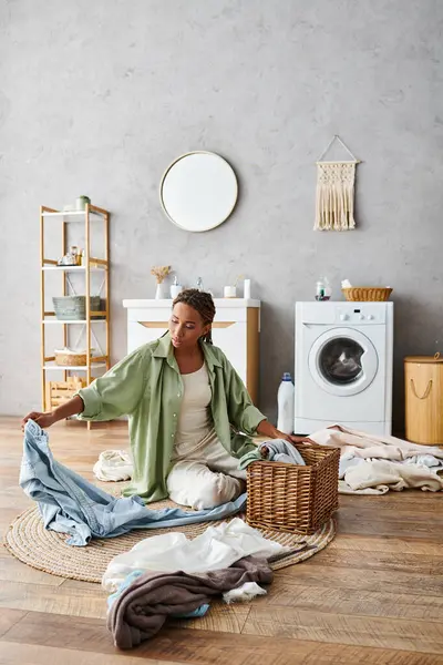 Eine Frau mit Afro-Zöpfen sitzt auf dem Boden vor einer Waschmaschine und wäscht ihre Wäsche im Badezimmer. — Stockfoto