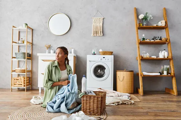 A woman with afro braids sits next to a washing machine, focused on doing laundry in a bathroom. — Stock Photo
