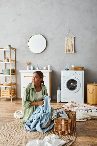 An African American woman with afro braids sitting on the floor in front of a washing machine, doing laundry in a bathroom. — Stock Photo