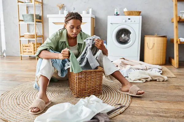 Eine afroamerikanische Frau mit Afro-Zöpfen sitzt auf dem Boden neben einem Wäschekorb, umgeben von Wäsche im Badezimmer. — Stockfoto